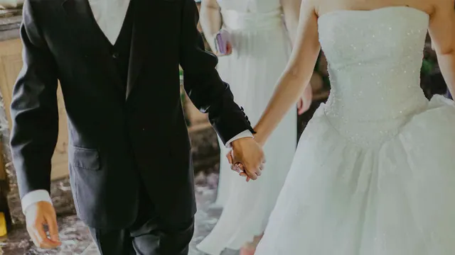 Illustrative photograph of a bride and groom holding hands, cropped below their faces.